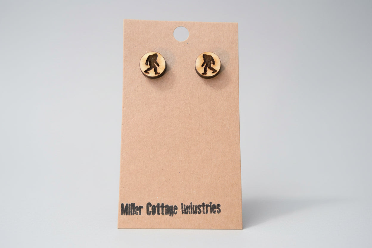 This image displays a pair of charming wooden stud earrings, crafted in the shape of a well-known Bigfoot silhouette. The earrings are meticulously designed with a textured, natural finish, enhancing their rustic appeal.